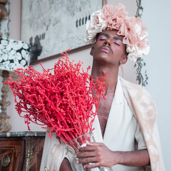 Ein schwarzer Mann trägt eine weiß-rosa Krone aus Blüten und hält einen Stauß korallfarbener Zweige in der Hand. Seine Augen sind geschlossen und sein Gesicht ist mit glitzernder Kosmetik verziert.