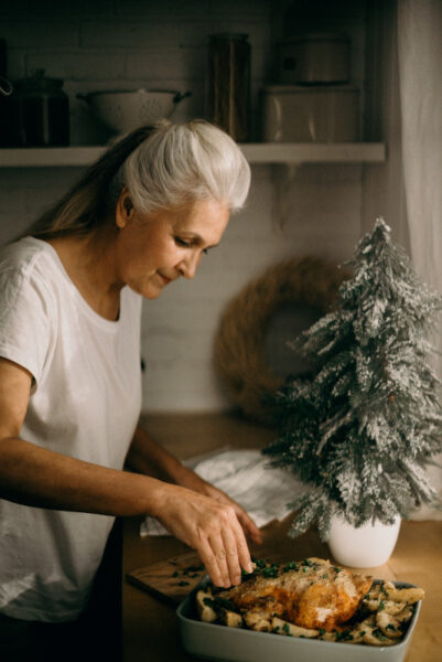 Eine ältere Dame garniert ein Weihnachtsmahl. Im Hintergrund steht ein Tannenbaum mit Glitzer-Kunstschnee verziert.