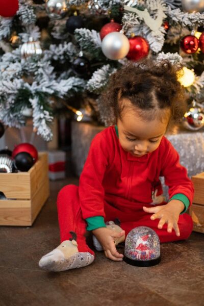Ein kleines Kind sitzt vor einem geschmückten Weihnachtsbaum und spielt mit einer Schneekugel