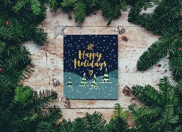 Zwischen einigen Tannenzweigen liegt eine Weihnachtskarte mit einem goldenen Schriftzug "Happy Holidays"