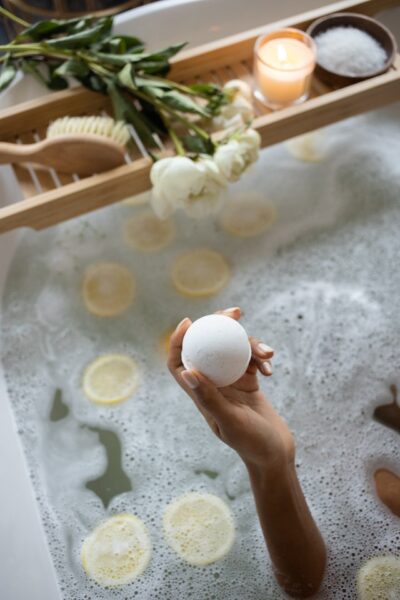 Eine Person sitzt in einer Badewanne, in der Zitronenschalen schwimmen und hält eine weiße Badekugel in der Hand