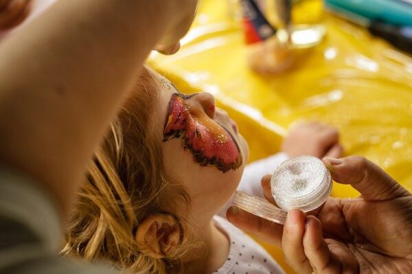 Einem Kind wird mit Glitzer und Farbe ein Schmetterling auf das Gesicht gemalt