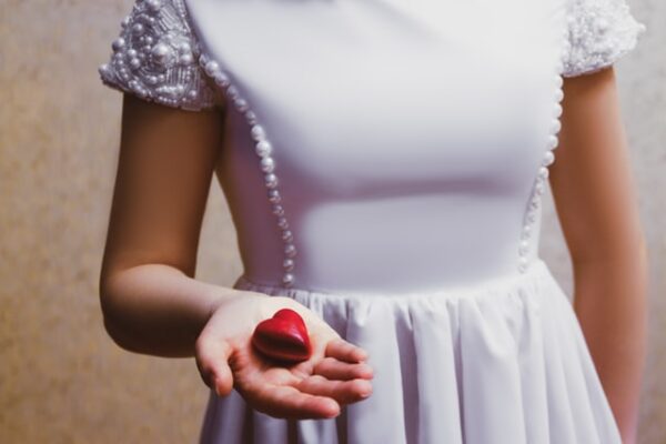Eine Frau mit weißem Kleid mit Perlen hält ein rotes Herz in der Hand